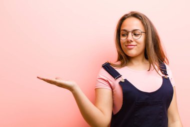 Genç bir kadın kendini mutlu hissediyor ve gelişigüzel gülümsüyor. Elinde pembe arka plana karşı duran bir nesneye ya da konsepte bakıyor.