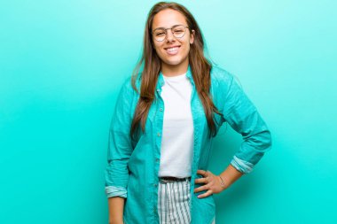 Genç bir kadın mutlu bir şekilde gülümsüyor, eli kalçasında ve kendinden emin, pozitif, gururlu ve arkadaş canlısı bir tavırla mavi arka plana karşı.