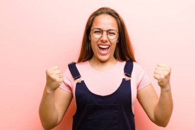 Genç bir kadın muzaffer bir şekilde bağırıyor, gülüyor, mutlu ve heyecanlı bir şekilde pembe arka plana karşı başarıyı kutluyor.