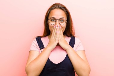 Genç kadın mutlu ve heyecanlı, şaşırmış ve hayrete düşmüş elleriyle ağzını kapatmış pembe arka plana karşı sevimli bir ifadeyle kıkırıyor.