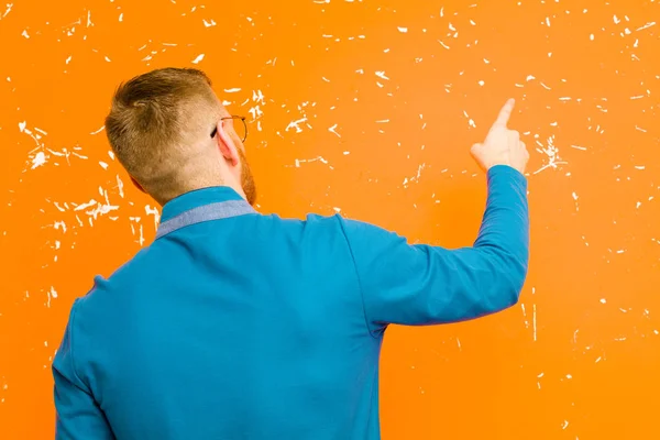若い赤の頭の男が立っているとコピースペースにオブジェクトを指して グランジオレンジの壁に対するリアビュー — ストック写真