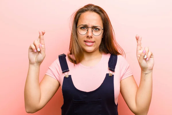 young  woman crossing fingers anxiously and hoping for good luck with a worried look against pink background