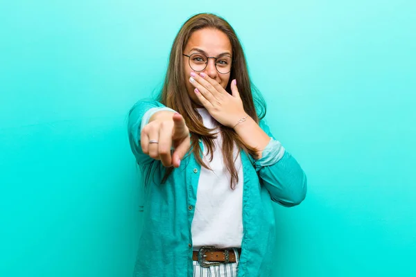 young woman laughing at you, pointing to camera and making fun of or mocking you against blue background