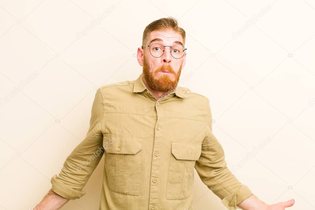 young red head man feeling clueless and confused, having no idea, absolutely puzzled with a dumb or foolish look against beige background