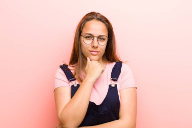 Genç bir kadın ciddi görünüyor, kafası karışık, kararsız ve düşünceli, pembe arka plana karşı seçenekler veya seçimler arasında şüpheci