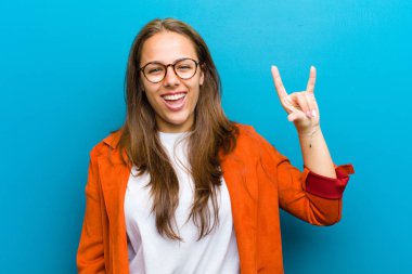 Genç bir kadın kendini mutlu, eğlenceli, kendinden emin, pozitif ve asi hissediyor, elleri mavi arka planda kaya ya da heavy metal işareti yapıyor.