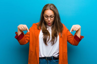 Genç bir kadın şoke olmuş, ağzı açık ve şaşırmış, mavi arka plana karşı inançsızlık ve şaşkınlık içinde bakıyor ve onu işaret ediyor.