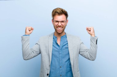 Genç işadamı mutlu, şaşırmış ve gururlu hissediyor, bağırıyor ve başarıyı kutluyor mavi arka planda büyük bir gülümsemeyle.