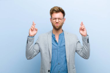 Genç işadamı gülümsüyor ve endişeyle parmaklarını çaprazlıyor, endişeli hissediyor ve mavi arka planda iyi şanslar diliyor veya umut ediyor.