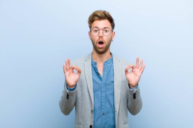 Genç işadamı şok olmuş, şaşırmış ve şaşırmış hissediyor. Onaylıyor. İki elini de mavi arka plana doğru uzatıyordu.