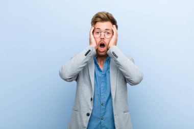 Genç işadamı mutsuz, korkmuş ya da endişeli görünüyor, ağzı sonuna kadar açık ve iki kulağını da mavi arka planda kapatıyor.