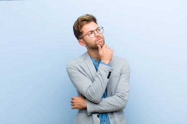 Genç işadamı düşünceli, meraklı ya da hayal kuruyor, hayal kuruyor ve mavi arka planda boşluğu kopyalamak için yukarı bakıyor.