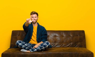 Pijamalı genç adam gülümsüyor ve arkadaş canlısı görünüyor, iki ya da iki numarayı gösteriyor, eli önde, geri sayıyor. Kanepede oturuyor.