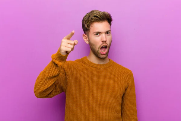 young man pointing at camera with an angry aggressive expression looking like a furious, crazy boss against purple background