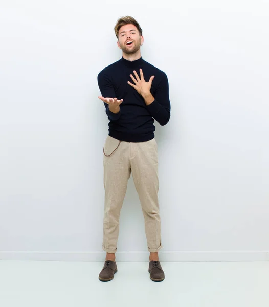 young full body man feeling happy and in love, smiling with one hand next to heart and the other stretched up front against white background