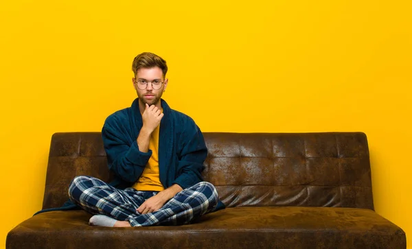 young man wearing pajamas looking serious, confused, uncertain and thoughtful, doubting among options or choices . sitting on a sofa