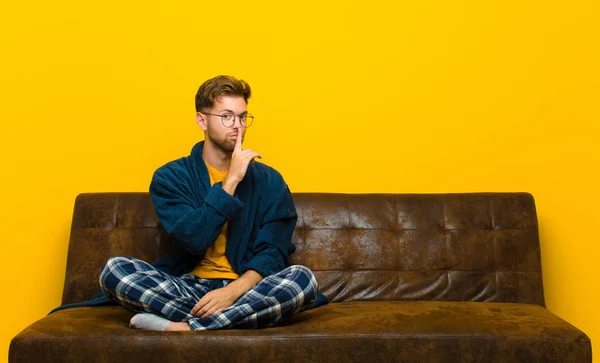young man wearing pajamas asking for silence and quiet, gesturing with finger in front of mouth, saying shh or keeping a secret . sitting on a sofa