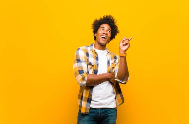 Genç siyah adam mutlu bir şekilde gülümsüyor ve yan bakıyor, merak ediyor, düşünüyor ya da turuncu duvara karşı bir fikri var.
