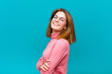 Genç ve güzel bir kadın kameraya çapraz kollarıyla gülümsüyor ve mutlu, kendinden emin, tatmin olmuş bir ifade, mavi arka planda yanal bir bakış.