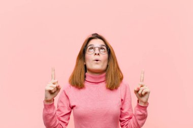 Genç ve güzel bir kadın huşu içinde ve ağzı açık bir şekilde pembe arka planda şaşkın ve şaşkın bir bakışla yukarıya bakıyor.