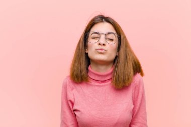 Genç, güzel bir kadın dudaklarını birbirine bastırıyor. Tatlı, eğlenceli, mutlu, sevimli bir yüz ifadesiyle. Pembe arka plana öpücük yolluyor.