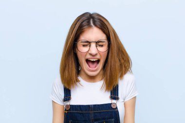 Genç ve güzel bir kadın agresif bir şekilde bağırıyor, sinirli, sinirli, öfkeli ya da sinirli görünüyor, mavi arka plana hayır diye bağırıyor.