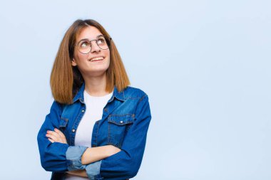 Genç, güzel bir kadın kendini mutlu, gururlu ve umutlu hissediyor, merak ediyor ya da düşünüyor, mavi arka planda çapraz kollarla fotokopi çekmeyi bekliyor.