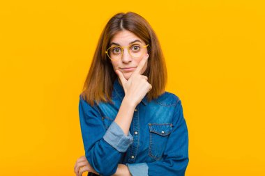 young pretty woman looking serious, thoughtful and distrustful, with one arm crossed and hand on chin, weighting options against yellow background clipart
