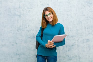 Genç bir bayan öğrenci utangaç ve neşeli bir şekilde gülüyor, arkadaşça ve olumlu ama güvensiz bir tavırla grunge duvarına karşı.