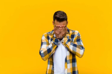 Genç İspanyol bir adam üzgün, hayal kırıklığına uğramış, gergin ve depresif hissediyor. İki eliyle yüzünü kapatıyor, turuncu duvara karşı ağlıyor.