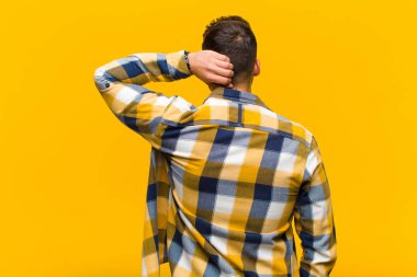 Genç İspanyol adam düşünüyor ya da şüphe ediyor, kafasını kaşıyor, şaşkın ve kafası karışık hissediyor, turuncu duvarın arkasında ya da arkasında.