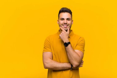 Genç İspanyol adam gülümsüyor, hayattan zevk alıyor, mutlu, arkadaş canlısı, tatmin olmuş ve elleri turuncu duvarda tasasız.
