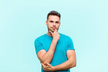 Genç İspanyol adam ciddi, şaşkın, kararsız ve düşünceli görünüyor. Mavi arka plandaki seçenekler veya tercihler arasında şüpheci.