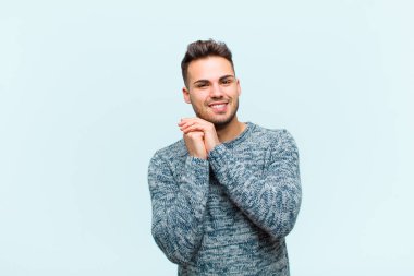 Genç İspanyol bir adam aşık ve sevimli görünüyor, sevimli ve mutlu, romantik bir şekilde gülümsüyor, elleri mavi arka planda.