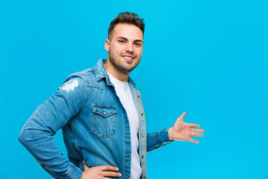 Genç İspanyol adam kendini mutlu ve neşeli hissediyor, gülümsüyor ve seni hoş karşılıyor, mavi arka plana karşı dostça bir jestle seni içeri davet ediyor.