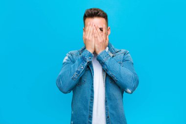 Genç İspanyol adam elleriyle yüzünü kapatıyor, parmaklarının arasından şaşkın bir ifadeyle bakıyor ve mavi arka plana bakıyor.