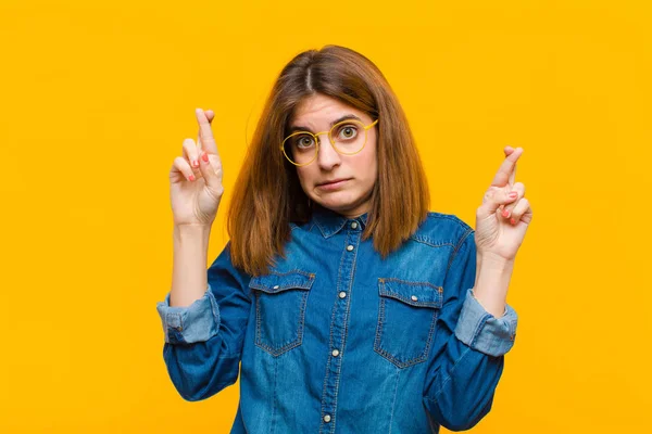 young pretty woman crossing fingers anxiously and hoping for good luck with a worried look against yellow background