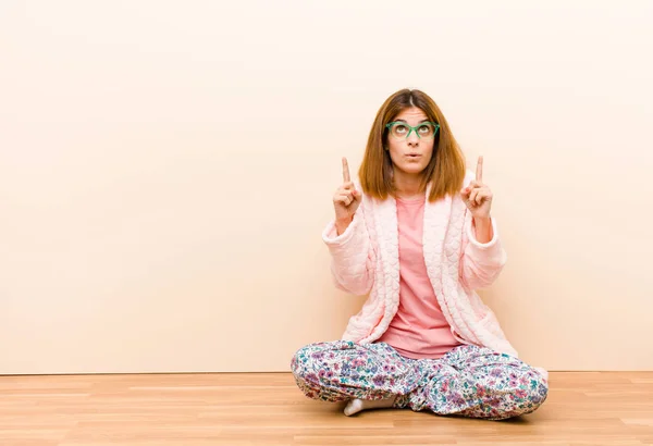 young woman wearing pajamas sitting at home feeling awed and open mouthed pointing upwards with a shocked and surprised look