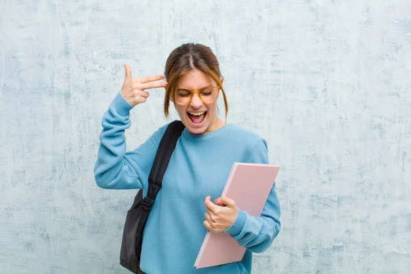 young student woman looking unhappy and stressed, suicide gesture making gun sign with hand, pointing to head against grunge wall background