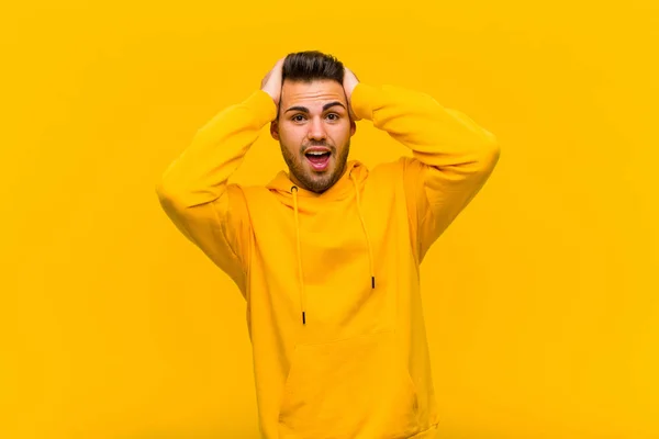 young hispanic man raising hands to head, open-mouthed, feeling extremely lucky, surprised, excited and happy against orange wall