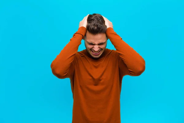 若いヒスパニック系の男はストレスと不満を感じ 頭に手を上げ 疲れて不満を感じ 青を背景に片頭痛で — ストック写真