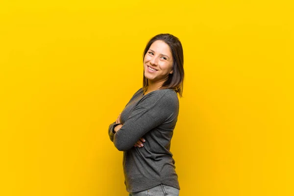 latin american woman smiling gleefully, feeling happy, satisfied and relaxed, with crossed arms and looking to the side isolated against yellow wall
