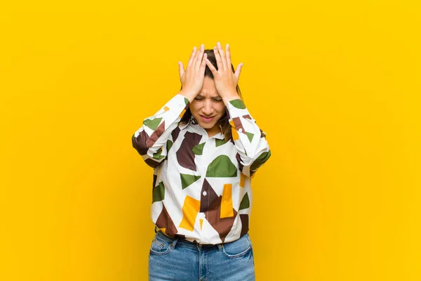 latin american woman feeling stressed and anxious, depressed and frustrated with a headache, raising both hands to head isolated against yellow wall