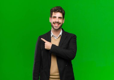 Genç işadamı neşeli bir şekilde gülümsüyor, mutlu hissediyor ve yan ve yukarı doğru işaret ediyor, fotokopi alanındaki nesneleri yeşil arka planda gösteriyor