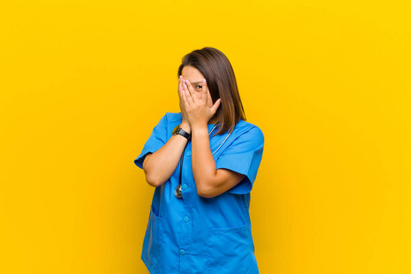 feeling scared or embarrassed, peeking or spying with eyes half-covered with hands isolated against yellow wall