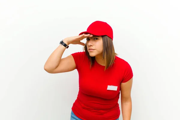 delivery woman looking bewildered and astonished, with hand over forehead looking far away, watching or searching against white background