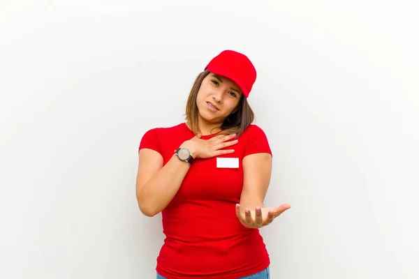 delivery woman feeling happy and in love, smiling with one hand next to heart and the other stretched up front against white background