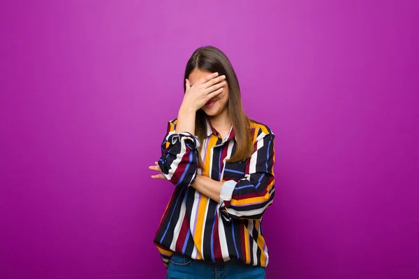 ストレス 恥ずかしい または動揺しているように見える若いかわいい女性は 頭痛で 紫色の背景に対して手で顔を覆う — ストック写真