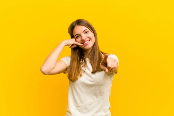 若い可愛い女性がカメラを指差しながら笑顔で後でジェスチャーをしてオレンジの背景に電話で話す — ストック写真