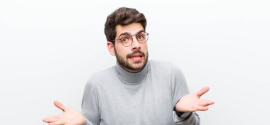 Genç yönetici şaşkın ve şaşkın hissediyor, şüpheci, farklı seçenekleri tartıyor veya beyaz duvara karşı komik bir ifadeyle seçiyor.